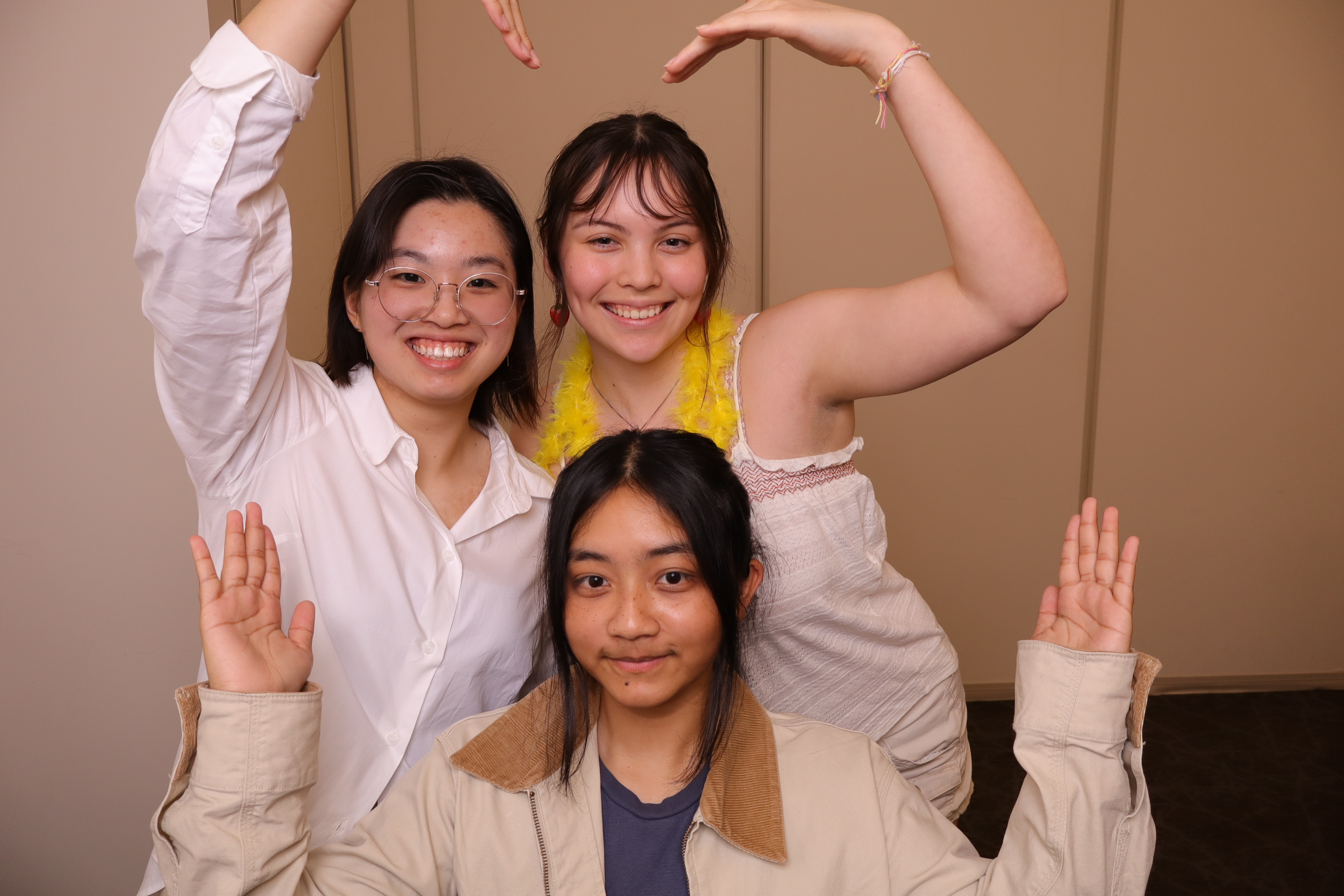 Three young women smiling, two behind forming a heart over the third's head, indoor photograph.