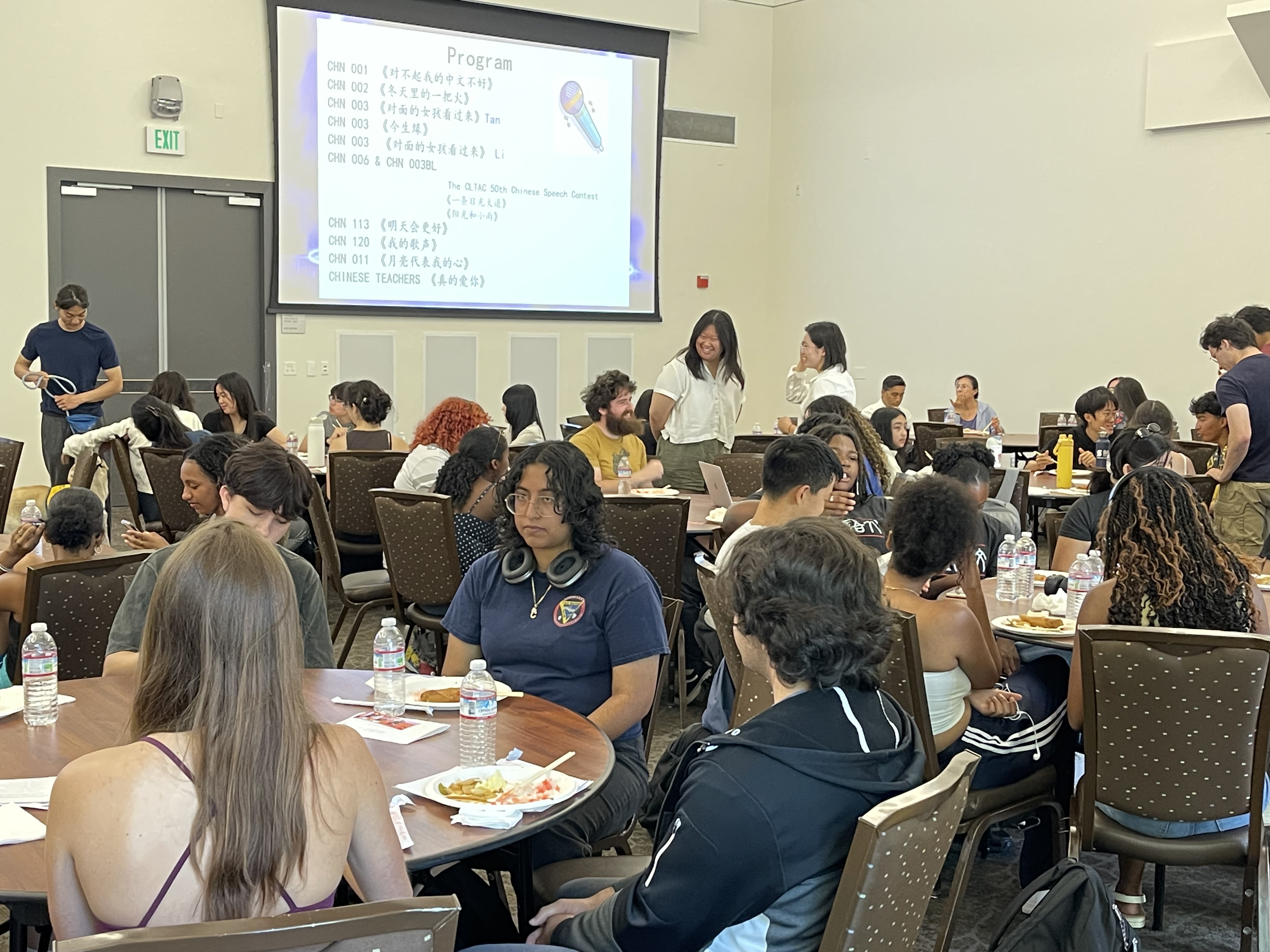 Conference room with diverse attendees seated at round tables, presenters and projected slide