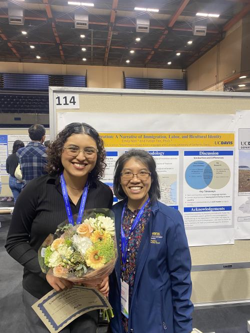 Two people stand near presentation, holding flowers and a certificate.